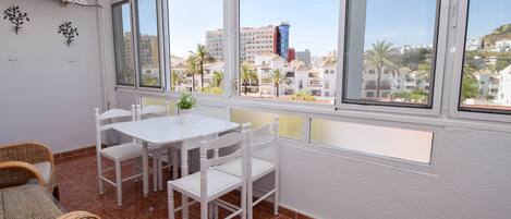 Een balkon met een witte tafel en stoelen, een rieten bank en uitzicht op gebouwen en palmbomen.