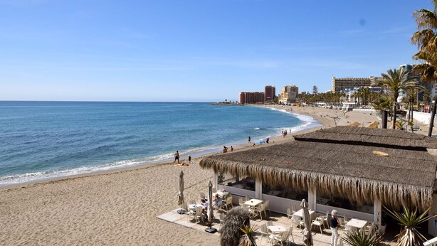 Un hotel frente al mar con un área de estar al aire libre con techo de paja, una playa arenosa y vistas al océano.