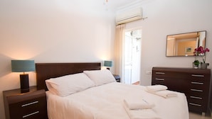 Una cama bien tendida con sábanas blancas, dos mesitas de noche con lámparas, un aparador con cajones y un espejo en la pared.