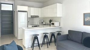 Eine moderne Küche mit weißen Schränken, einer eingebauten Mikrowelle und einer Kaffeemaschine. Im Wohnbereich befinden sich drei schwarze Hocker und ein graues Sofa.