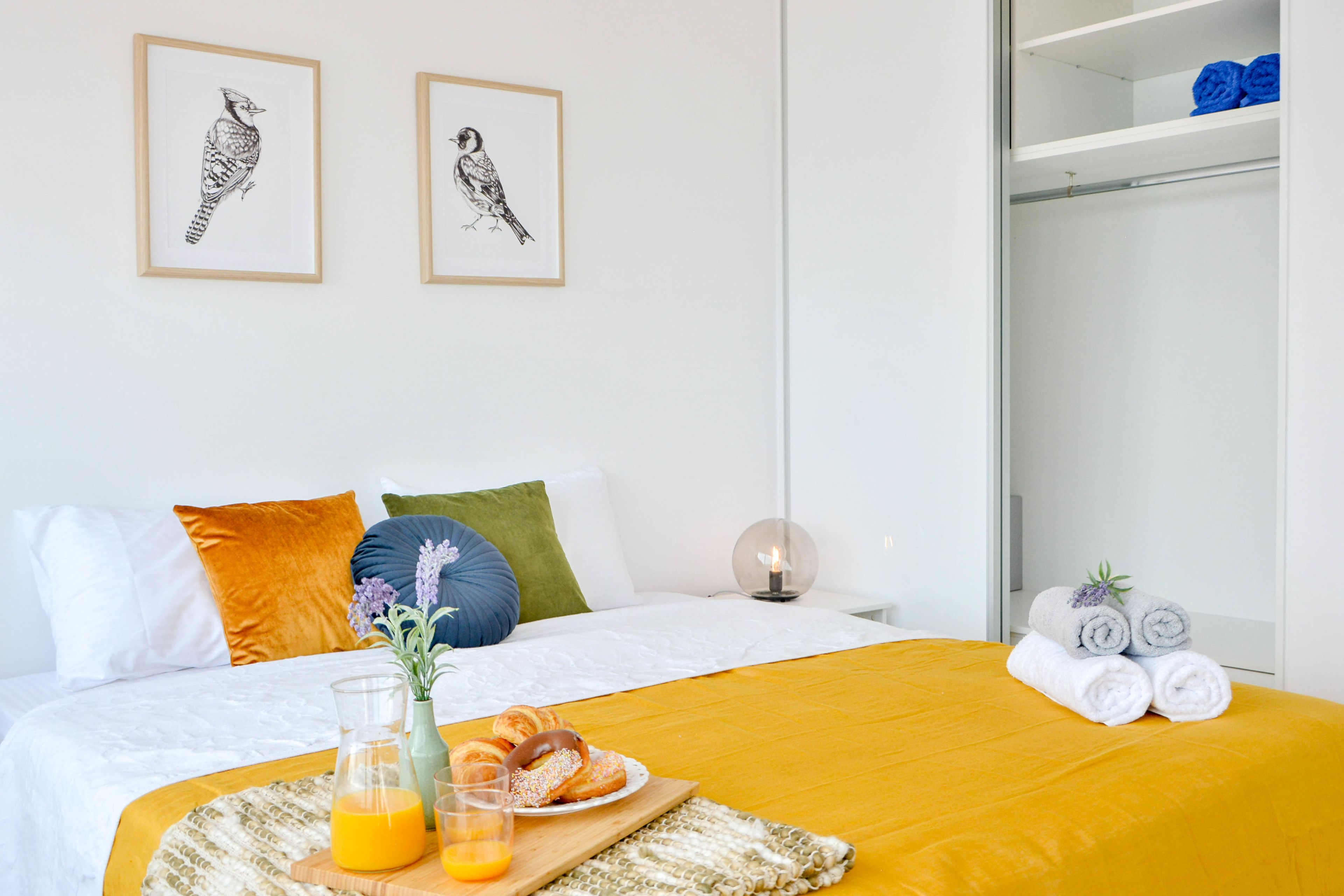 A neatly made bed with a yellow bedspread, a bedside table with a glass of orange juice, and a plate of croissants. There are two framed bird illustrations on the wall.