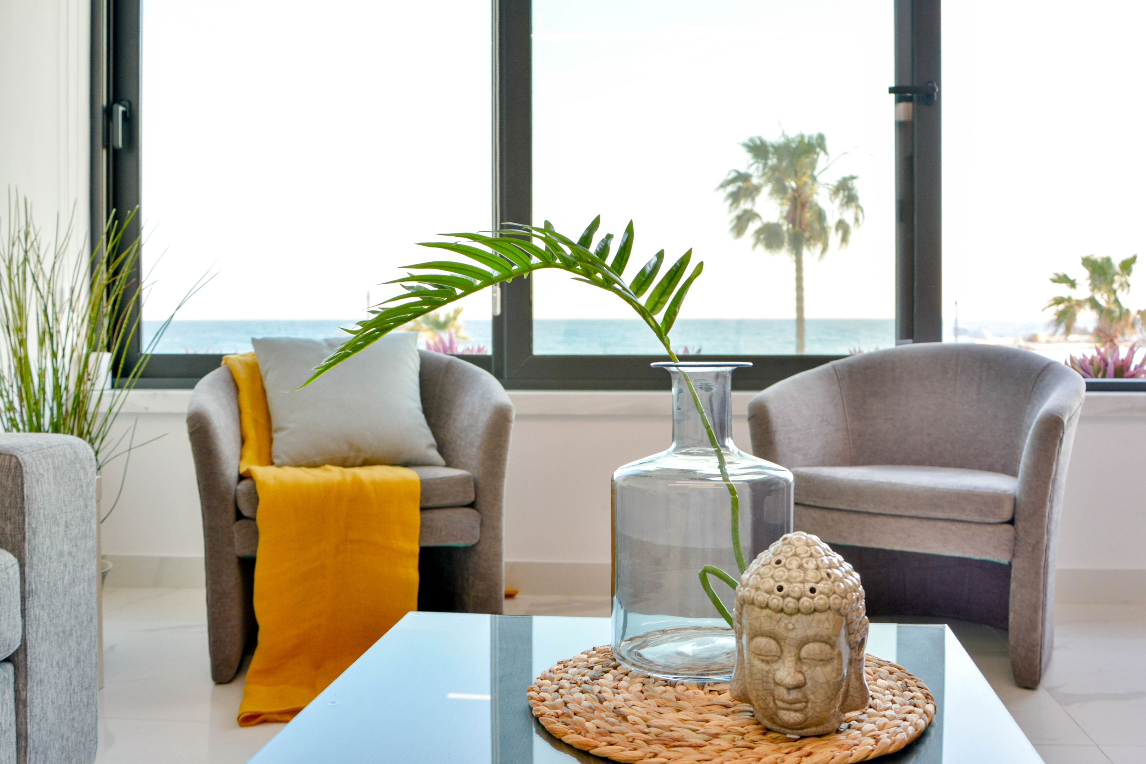 En moderne stue med havudsigt, med to grå lænestole, en glasvase med en plante og en Buddha-statue på en vævet bakke.