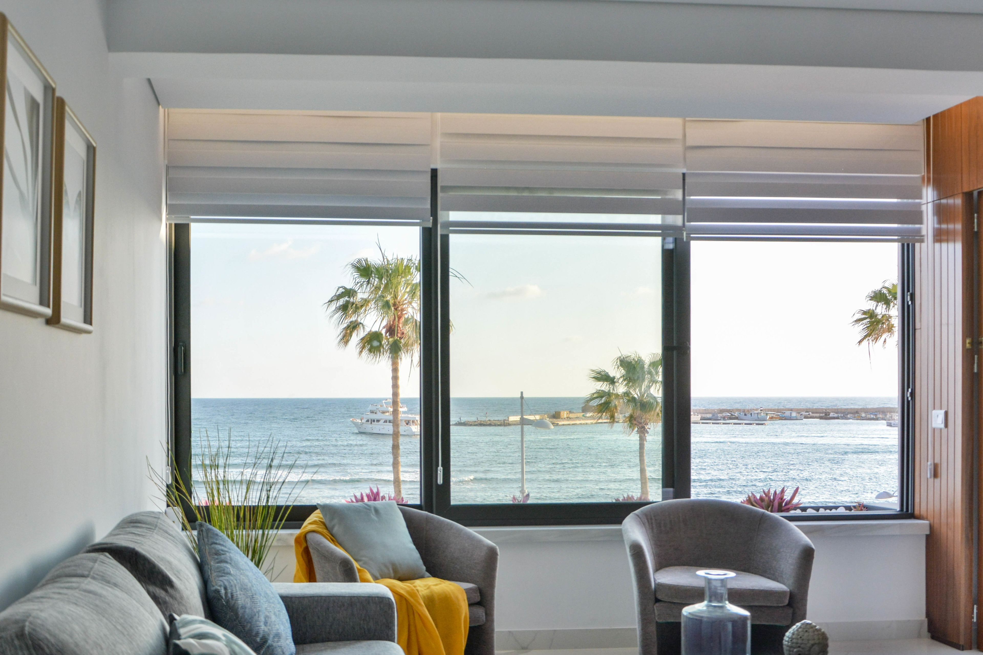 A room with a large window offering a view of the sea and palm trees. There are two armchairs and a sofa with cushions.