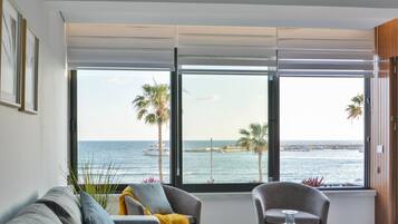 A room with a large window offering a view of the sea and palm trees. There are two armchairs and a sofa with cushions.