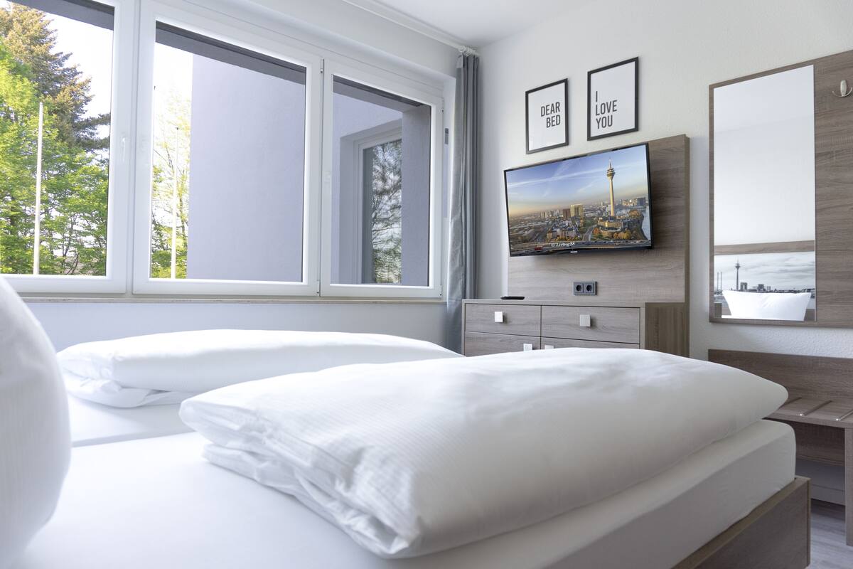 A hotel room with two beds, a TV mounted on the wall, and a window with a view of trees.