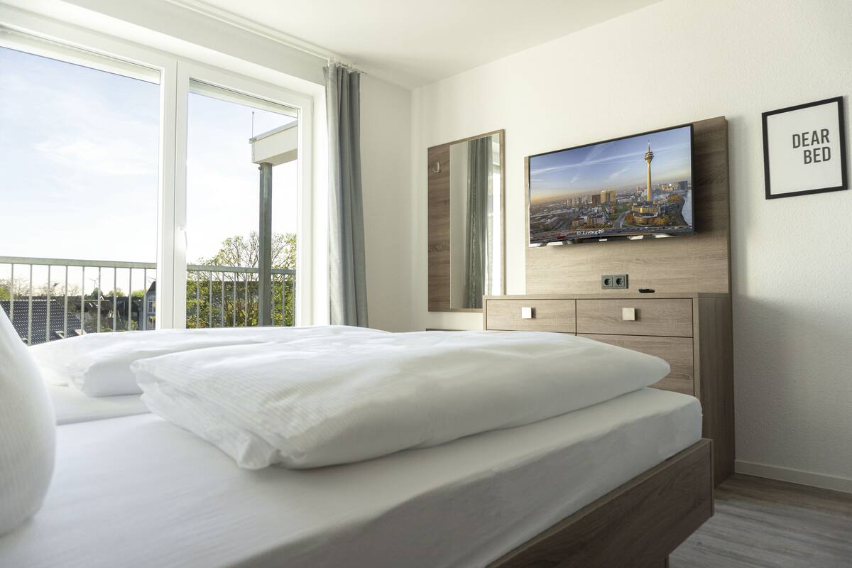 A hotel room with a large bed, a TV mounted on the wall, a mirror, and a view of a cityscape through the window.
