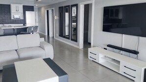 A modern living room with a sofa, a coffee table, a TV, and a kitchen area with a microwave and cabinets. - 504 Coral Reef (La Mercy)
