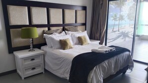 A hotel room with a large bed, a nightstand, a lamp, and a view of a beach through the window. - 504 Coral Reef (La Mercy)