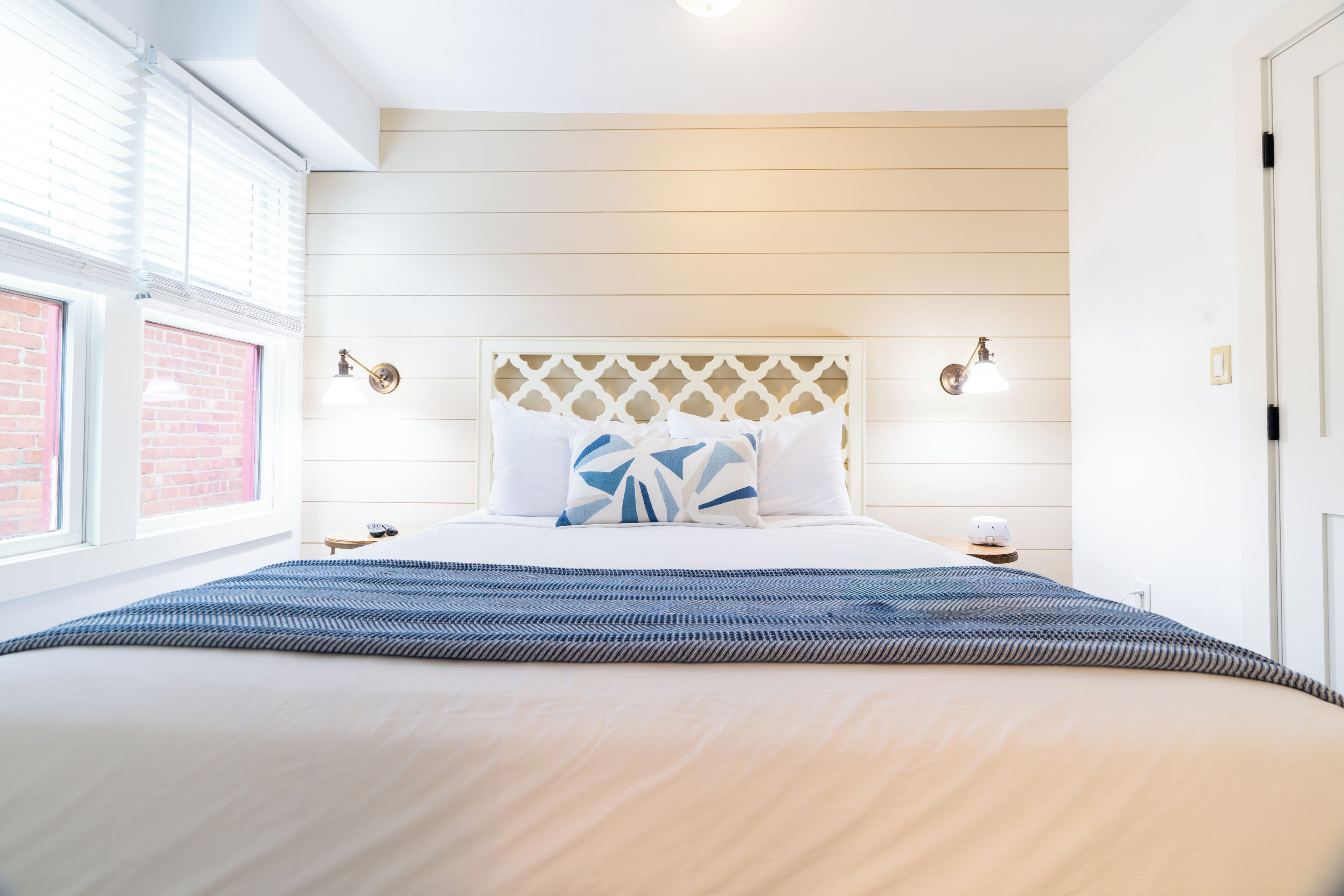 A neatly arranged bedroom with a large bed, a headboard with a lattice design, two bedside lamps, and a window with blinds.