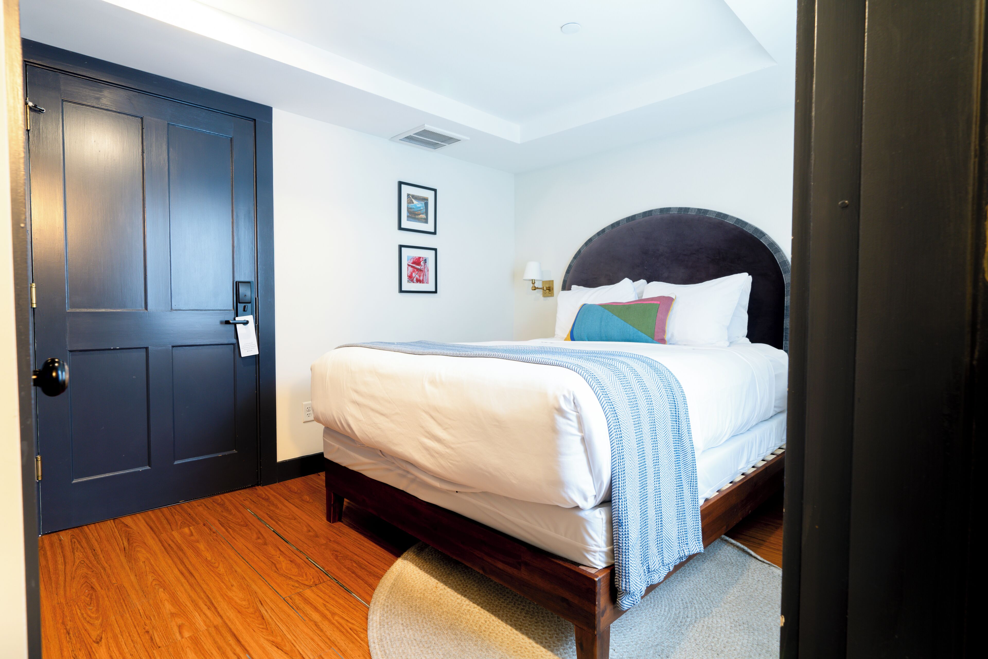 A bedroom with a dark wooden door, a bed with white bedding and a blue and white striped blanket, two framed pictures on the wall, and a small rug on the floor.