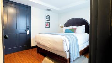 Quarto com porta de madeira escura, cama com roupa de cama branca e manta listrada em azul e branco, duas fotos emolduradas na parede e um tapete pequeno no chão.