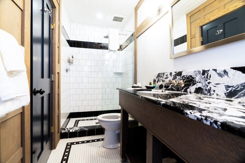 A modern bathroom with a black and white tiled floor, a white toilet, a black marble sink, and a wooden door.
