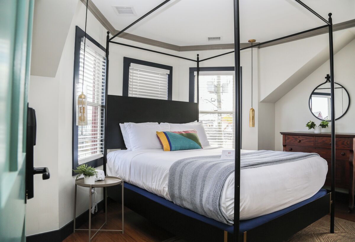 A four-poster bed with a black frame, white bedding, and a blue mattress. There is a small bedside table with a plant and a lamp. The room has a sloped ceiling and a window with blinds.