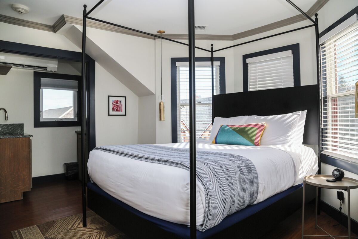 A four-poster bed with a white and blue bedspread, a small bedside table with a lamp, and a window with blinds.