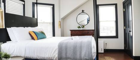 A bedroom with a four-poster bed, a wooden dresser, a mirror, and a view of the city through large windows.