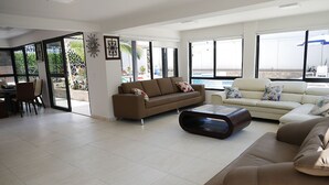Een ruime woonkamer met een bruine bank, een witte bank en een salontafel. Er is een glazen deur die leidt naar een terras met een zwembad.