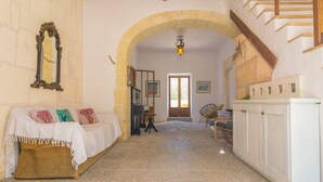 A room with a stone wall, a wooden staircase, a white sofa with colorful cushions, a white cabinet, and a door leading outside. - SON BANUS - Chalet with private garden in Manacor. Free WiFi (Manacor)