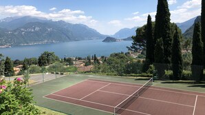 Een tennisbaan met een net, omringd door groen en een meer met bergen op de achtergrond.