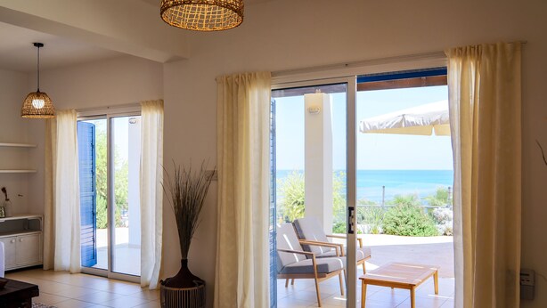 A spacious room with a large sliding glass door leading to a balcony with a view of the sea. There is a seating area with a table and chairs, and a potted plant.