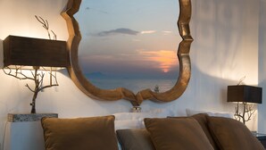 A bedroom with a large mirror reflecting a sunset, a bed with multiple pillows, and two bedside lamps. - Villa Rufola (Sorrento)