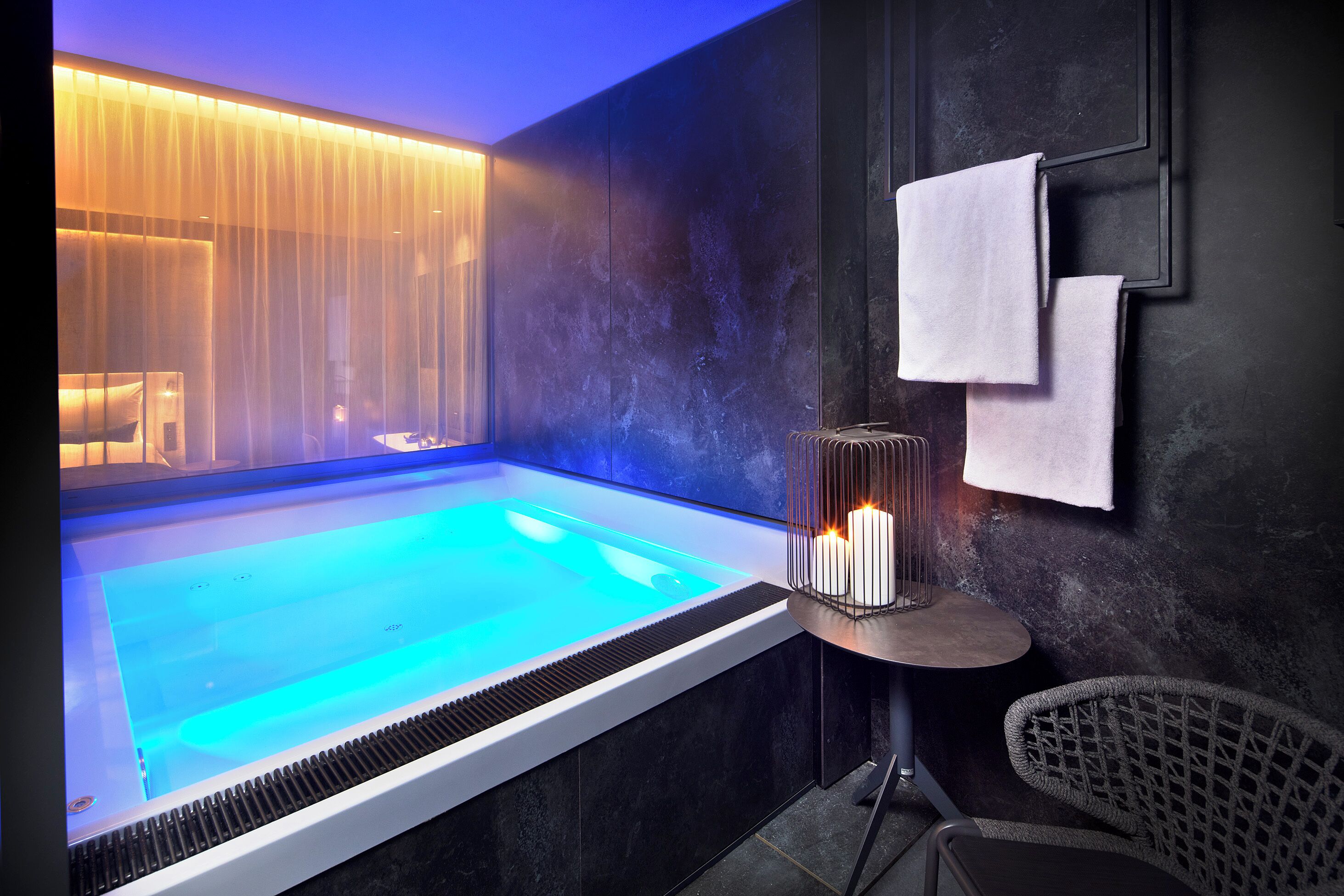 A modern spa room with a heated pool, a small table with candles, and a chair.