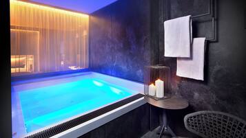 A modern spa room with a heated pool, a small table with candles, and a chair.