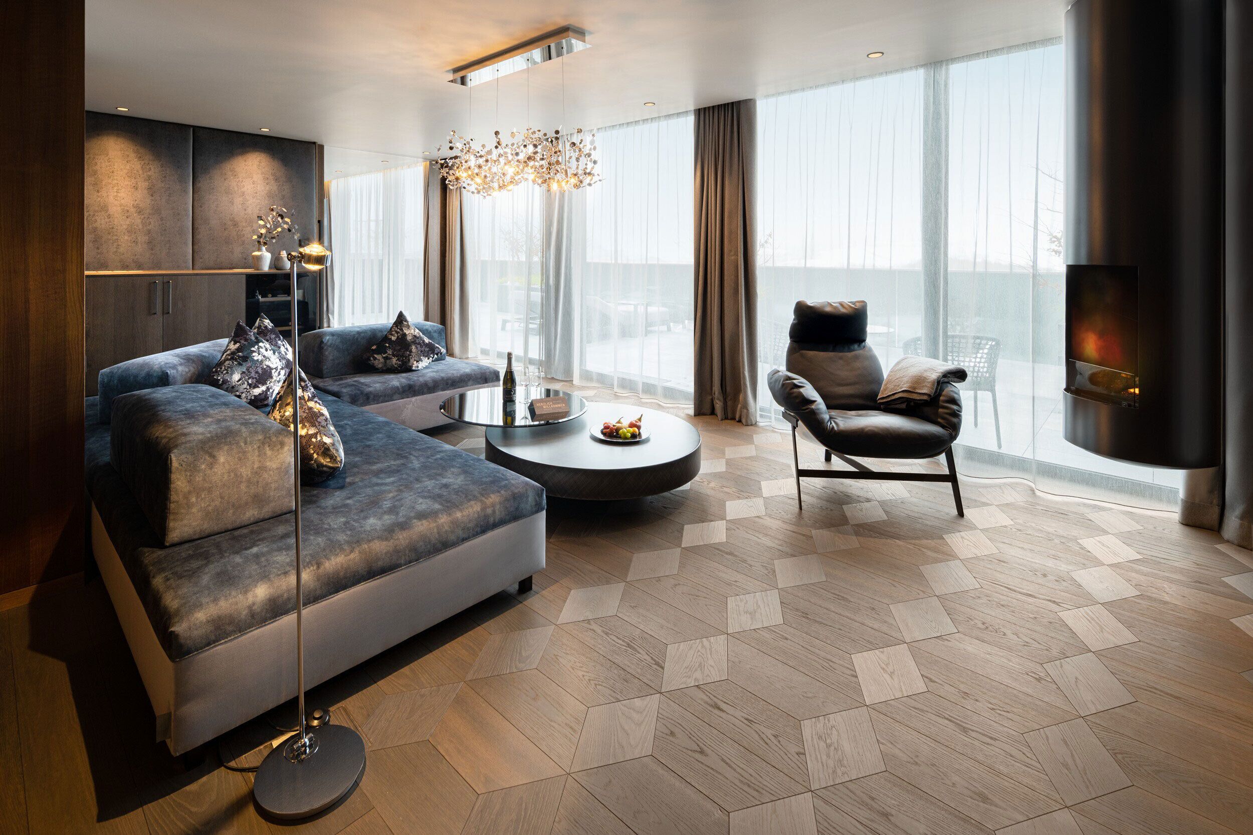 A modern living room with a sectional sofa, a round coffee table, a leather armchair, and a large window offering a view of the city.
