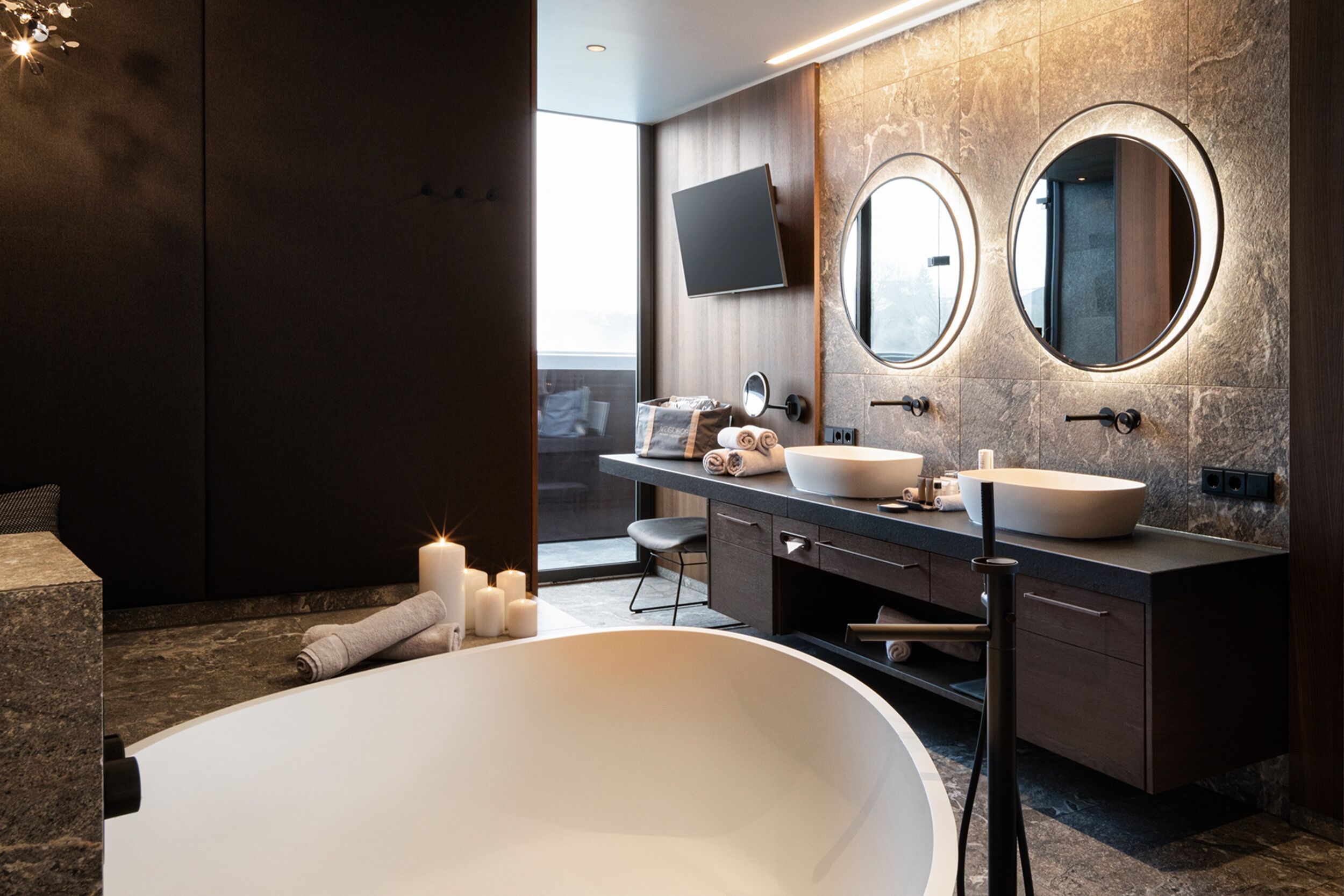 Casa de banho moderna com uma banheira independente, dois espelhos redondos, uma pia grande e uma televisão de ecrã plano fixada na parede.