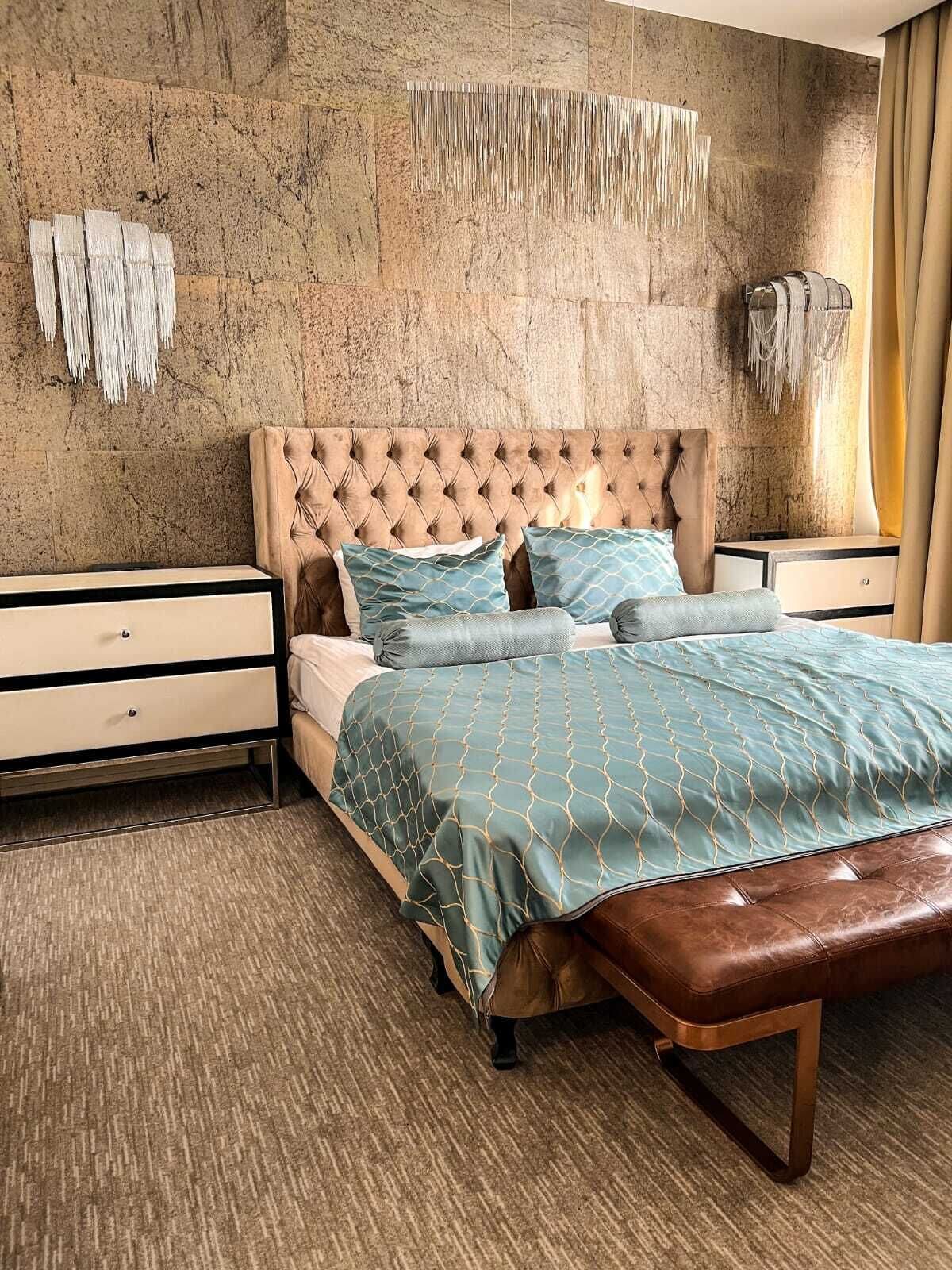 A bedroom with a tufted headboard, a bed with a patterned bedspread, two bedside tables with drawers, and a bench at the foot of the bed.