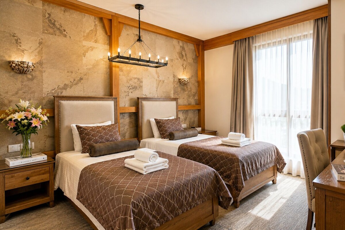 A hotel room with two beds, a wooden headboard, a chandelier, a painting, and a desk with a lamp and a book.