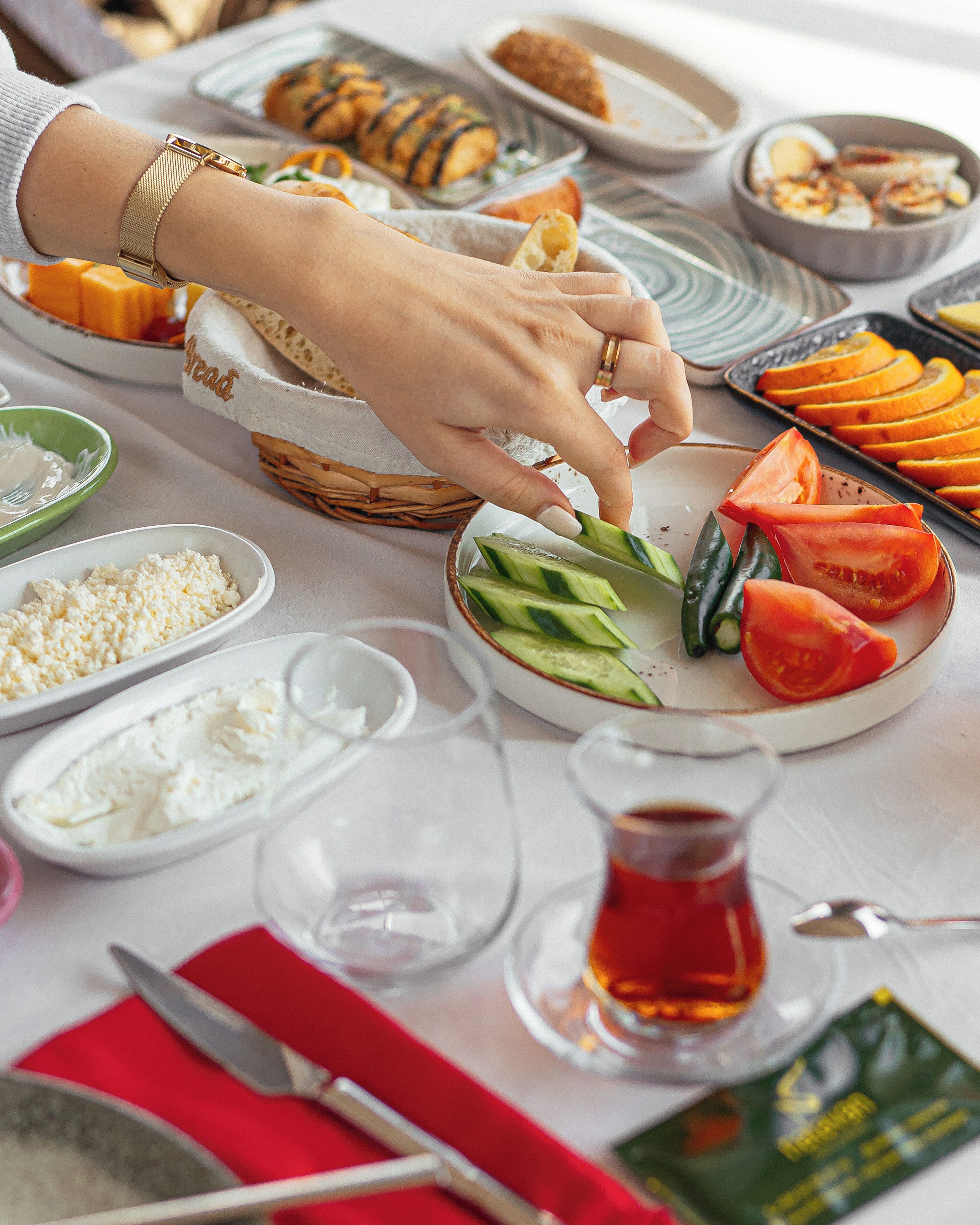 Ein Tisch ist reichhaltig mit Speisen gedeckt, darunter Tomatenscheiben, Gurken und Käse. Eine Person richtet das Essen her.