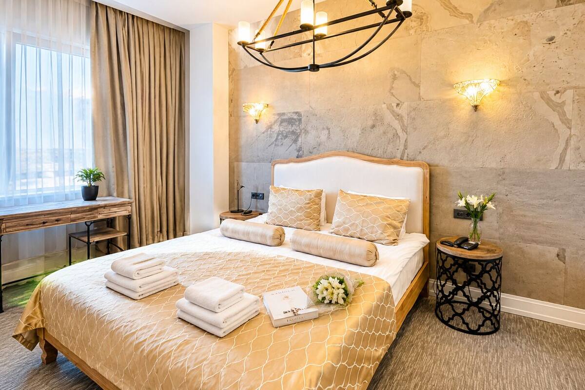 Quarto de hotel com cama, escrivaninha, cadeira, mesa de cabeceira e luminária fixada na parede.