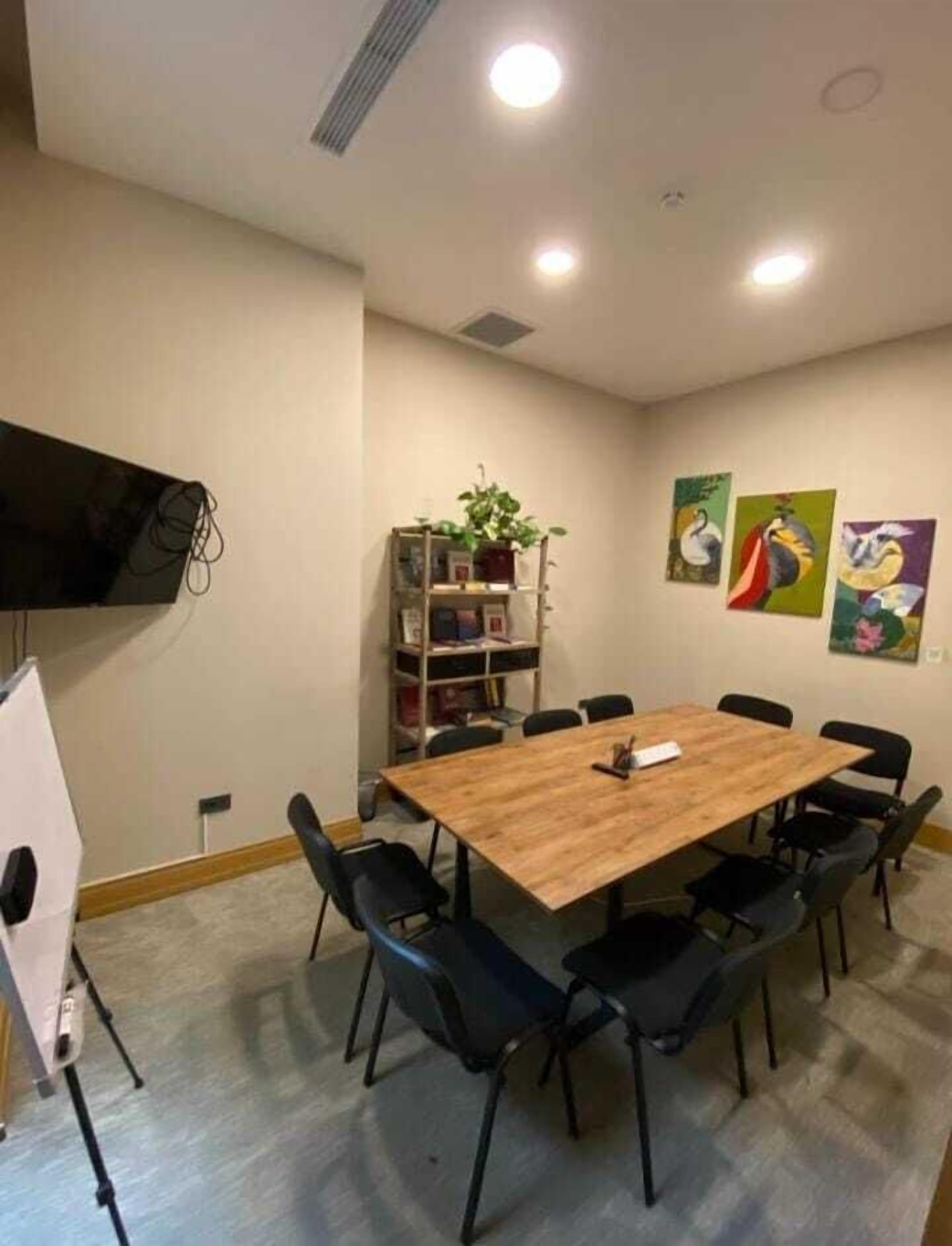 A conference room with a wooden table, black chairs, a bookshelf, and three paintings on the wall.