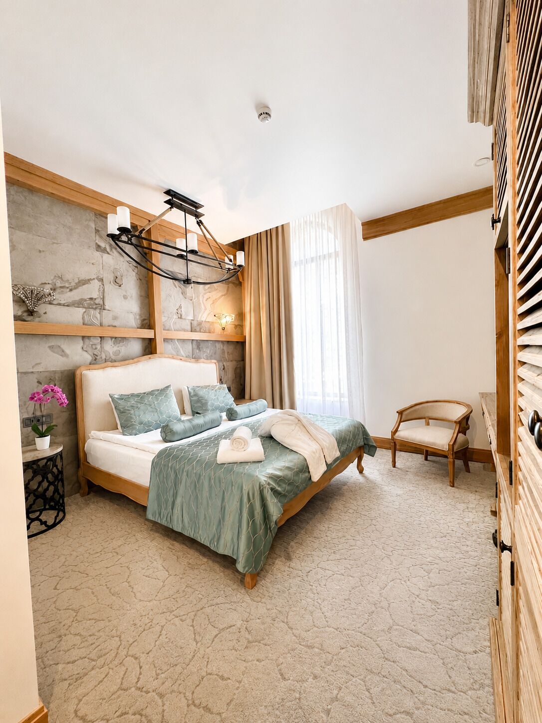 A bedroom with a bed, a nightstand, a chair, a lamp, and a window with curtains.