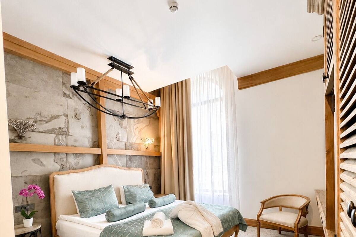 Quarto com cama, mesa de cabeceira, cadeira, luminária e janela com cortinas.