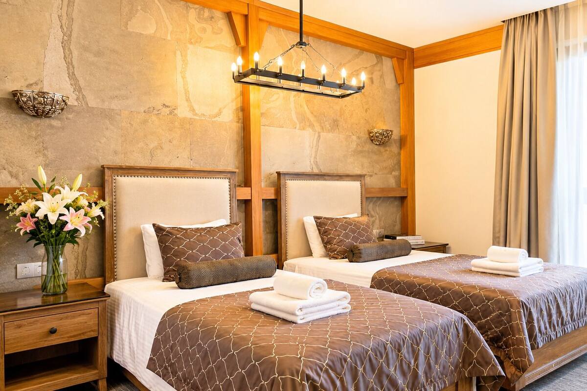 Quarto de hotel com duas camas, parede de pedra, lustre e janela com cortinas.