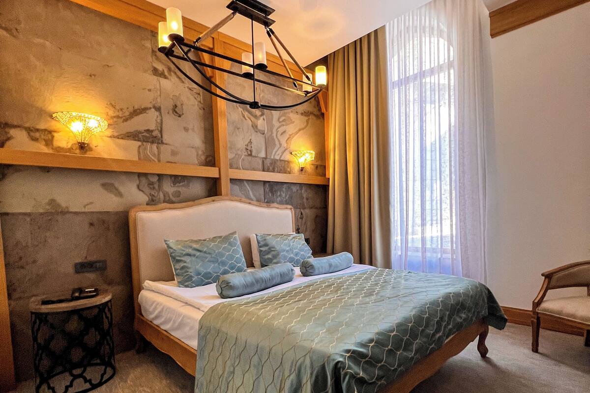 Quarto com cama, mesa de cabeceira, cadeira, lustre e janela com cortinas.