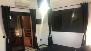 Una habitación con cama, una mesita con lámpara, una ventana con cortinas y una puerta corredera que da a otra habitación.