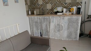 Et opholdsområde med en grå sofa, et lille flet kaffebord med en orkidé i potte, samt et køkkenområde med mønstrede fliser og en kaffemaskine.