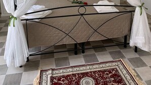 En seng med baldakin og fire søjler, med en quiltet madras, hvide puder og et rødt og hvidt tæppe på et flisegulv.