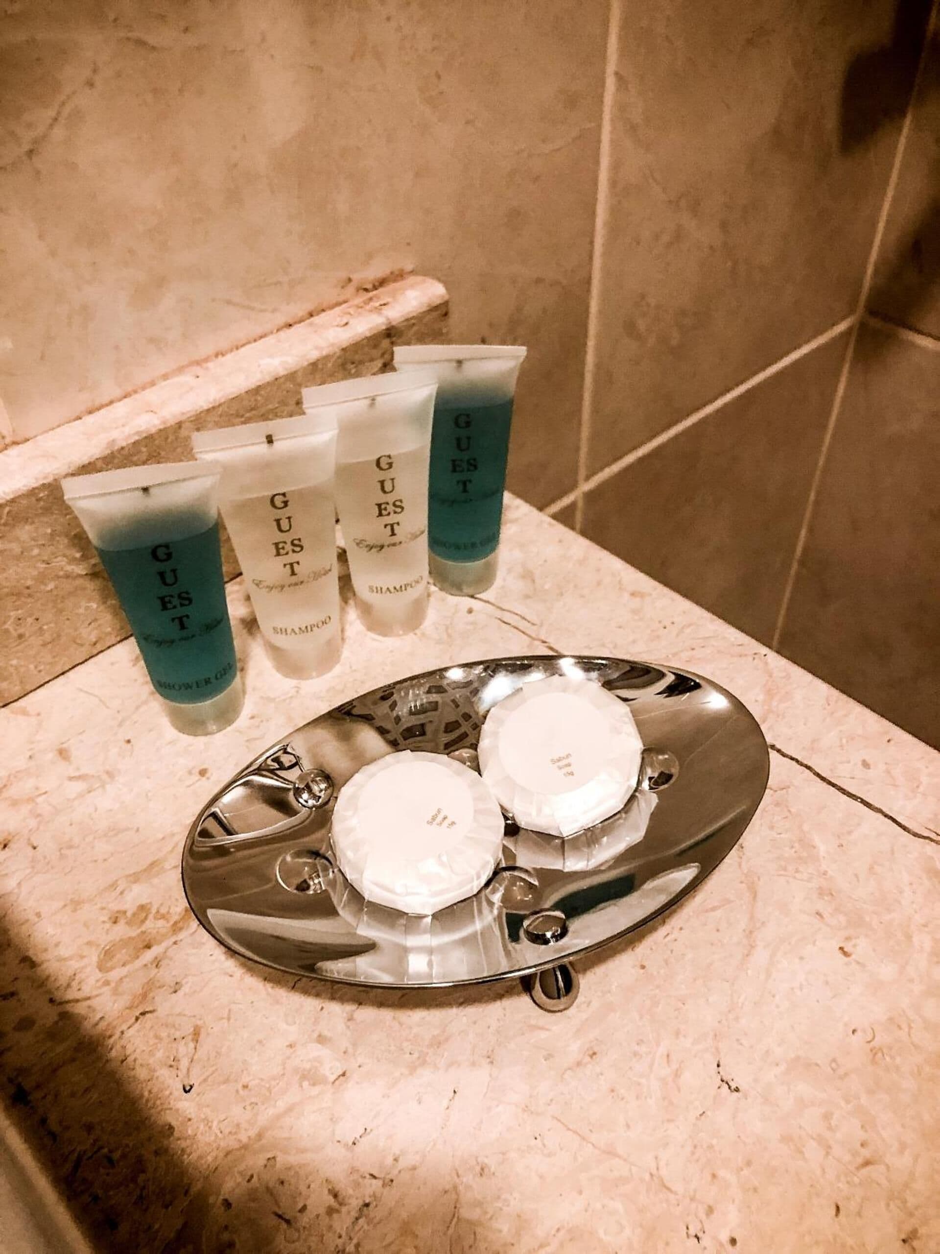 A bathroom sink with four tubes of guest shampoo and conditioner, and two white shower caps on a silver tray.