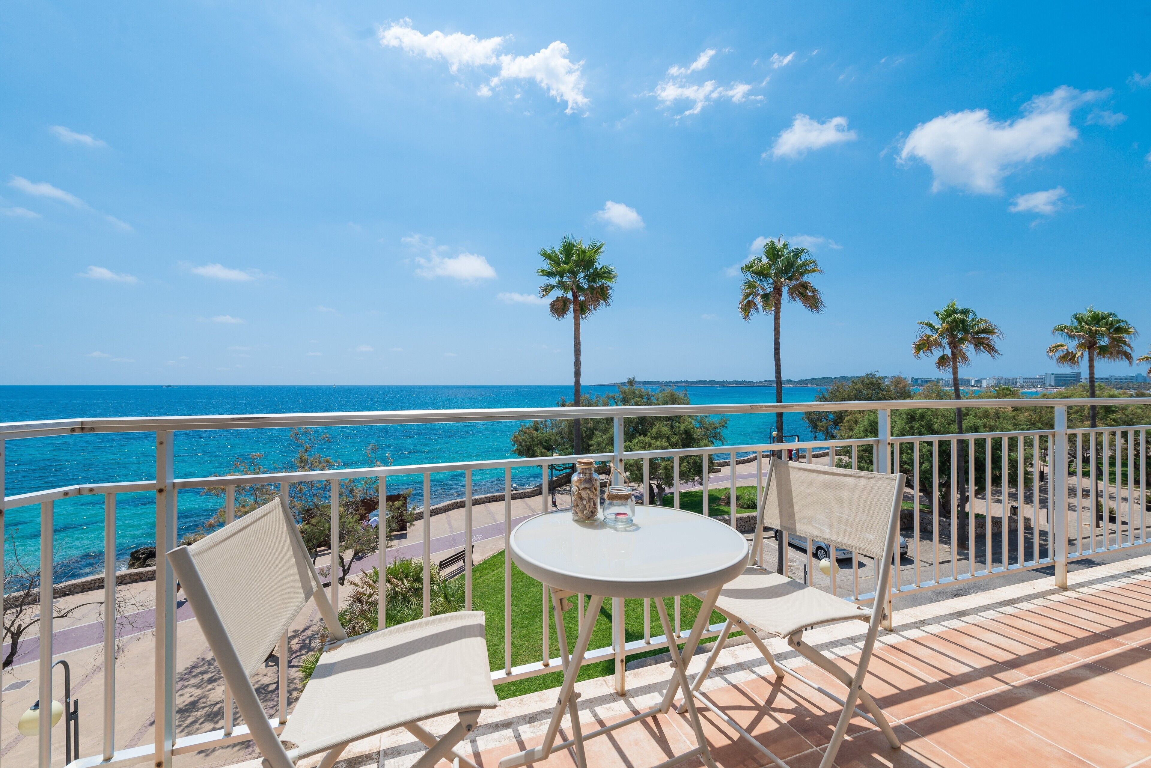 Un balcón con dos sillas y una mesa, con vista a una playa con palmeras y el mar.