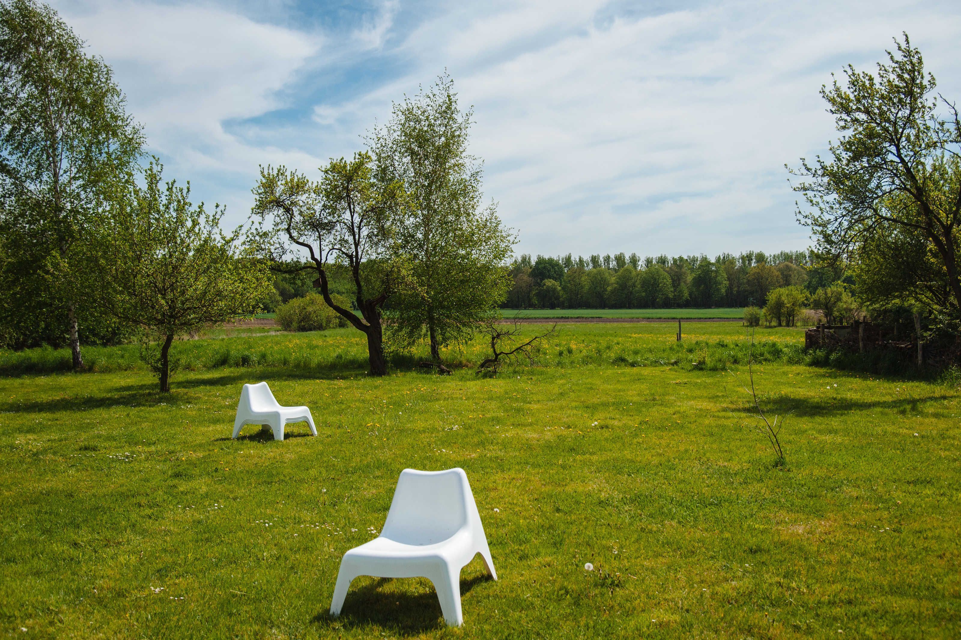 Zwei weiße Plastikstühle in einer grünen Wiese mit Bäumen und blauem Himmel.