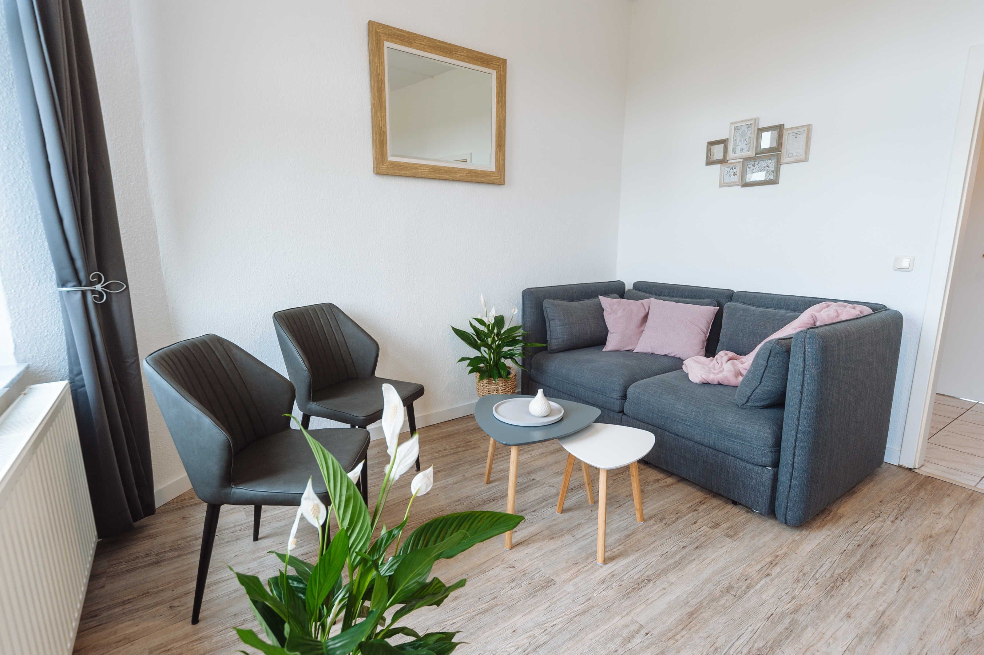 Ein modernes Wohnzimmer mit einem grauen Sofa, zwei Sesseln, einem kleinen Couchtisch und einer Topfpflanze.