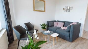 Ein modernes Wohnzimmer mit einem grauen Sofa, zwei Sesseln, einem kleinen Couchtisch und einer Topfpflanze.