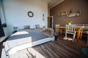 Un dormitorio con una cama, una mesa, sillas, una lámpara de mesa pequeña, un estante en la pared con objetos decorativos y una puerta.