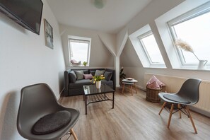 Ein modernes Wohnzimmer mit einer Couch, einem Glastisch und einem Sessel. Es gibt zwei Fenster und eine Heizung.