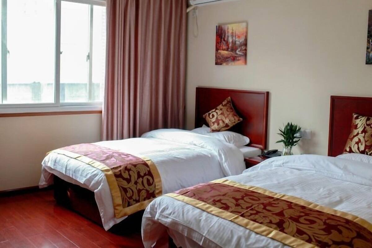 A hotel room with two beds, a window with curtains, an air conditioner, and a painting on the wall.