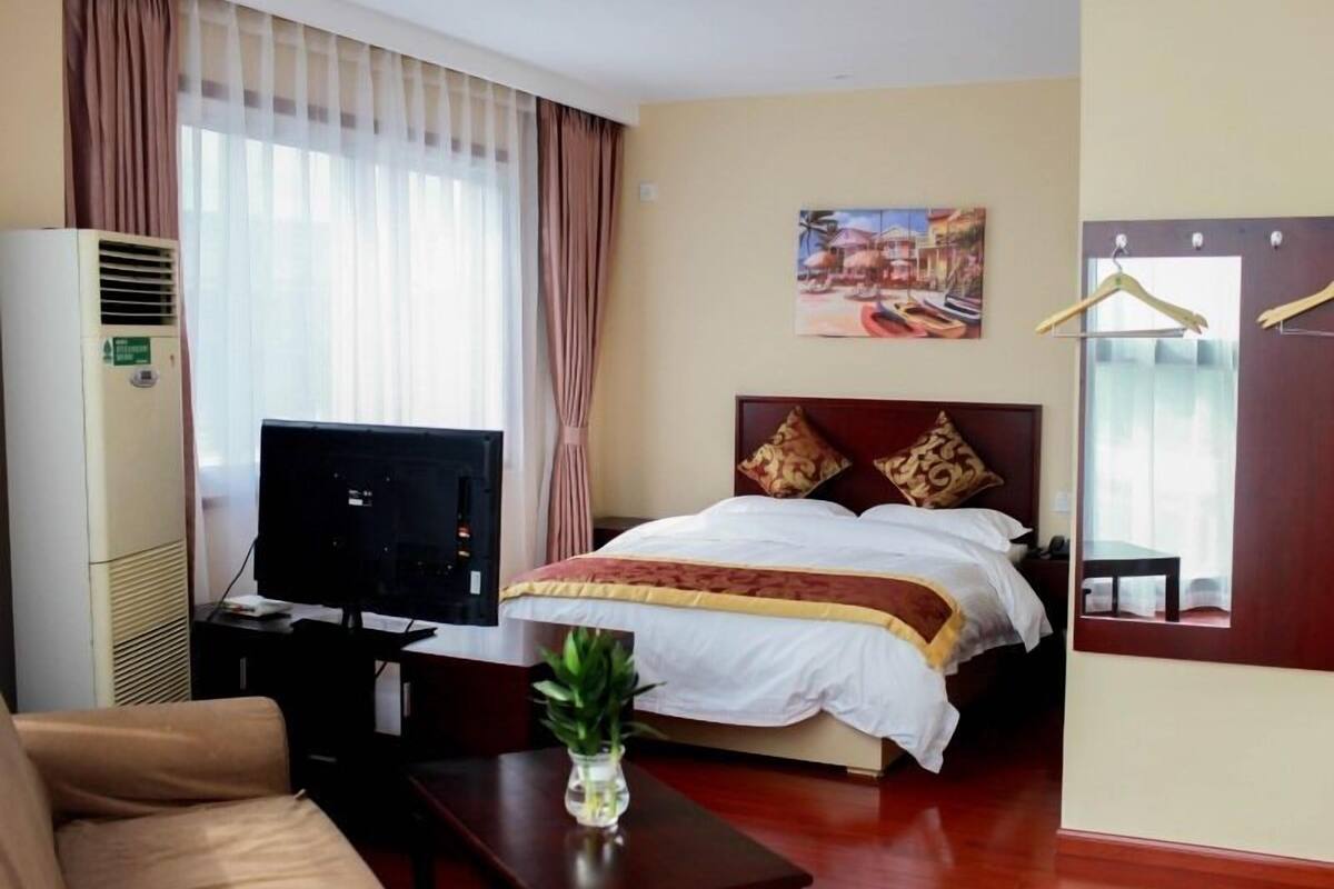 A hotel room with a bed, a TV, a desk, a chair, a plant, and a painting on the wall.
