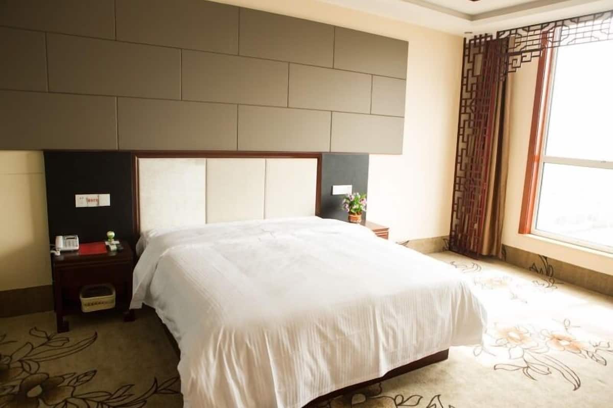 Une chambre d’hôtel avec un grand lit, une tête de lit rembourrée, une table de chevet avec un téléphone et une petite plante, et une fenêtre avec des rideaux.
